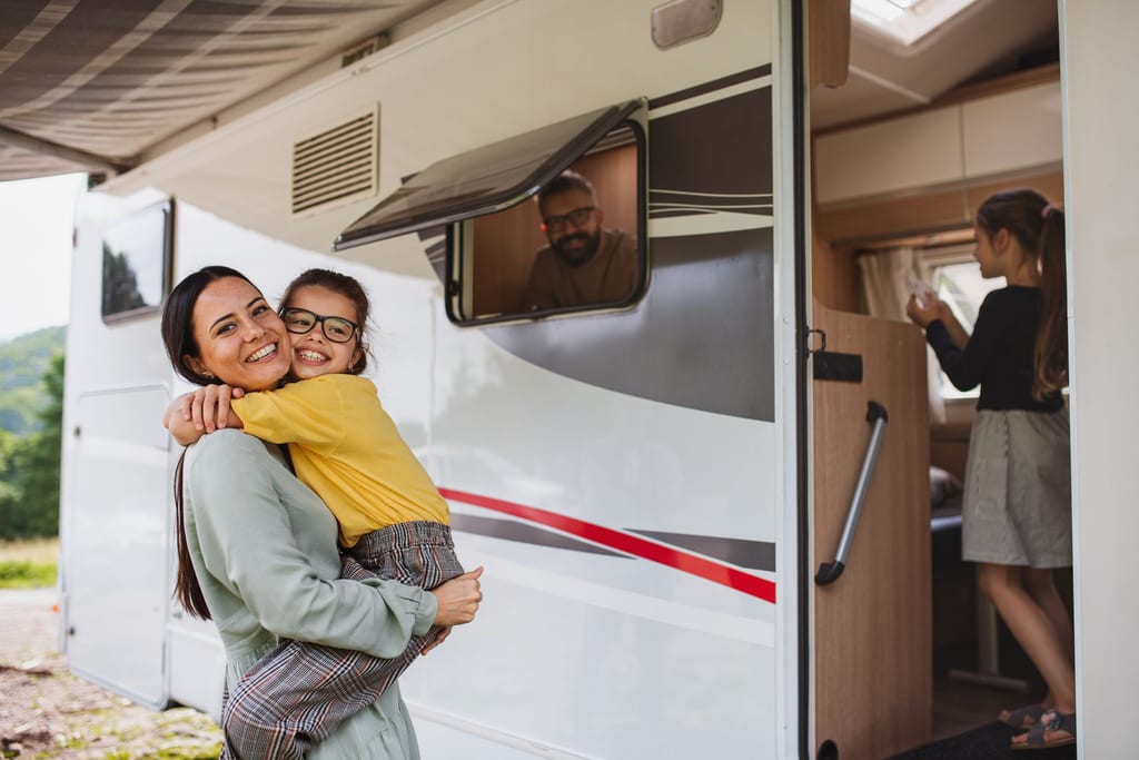 A mother and daughter hugging outside the open door of their travel trailer, with family inside.
