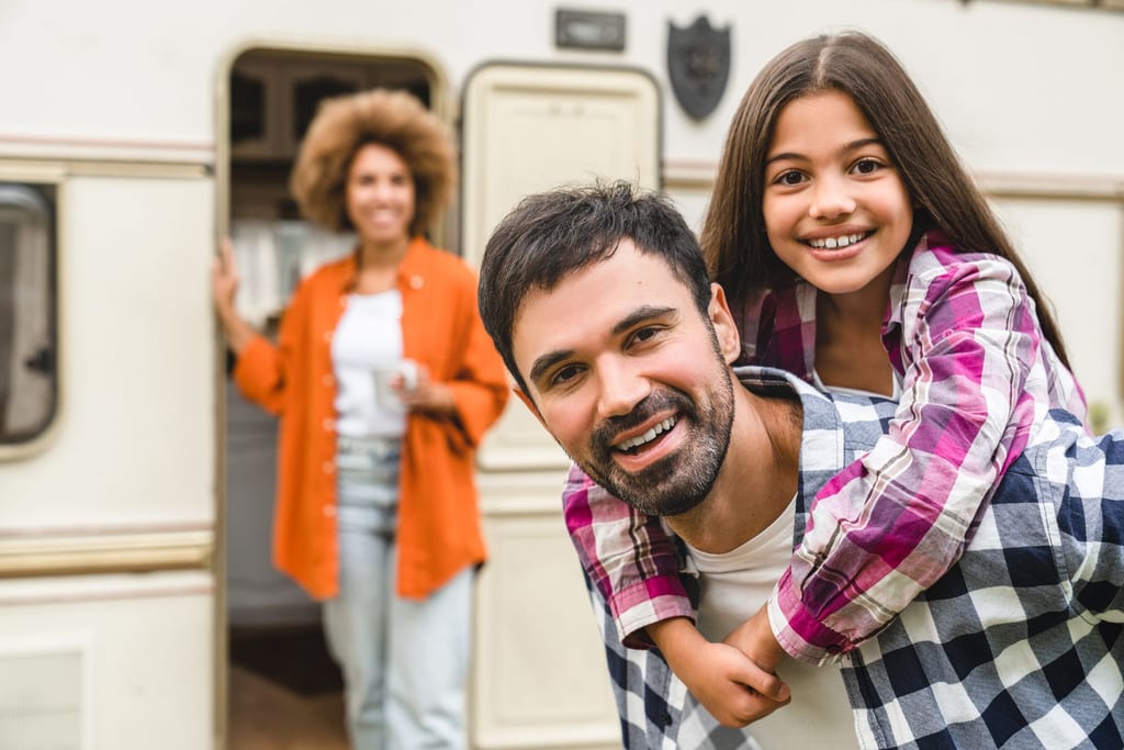 Smiling man giving a girl a piggyback near an open camper, with a woman standing in the background