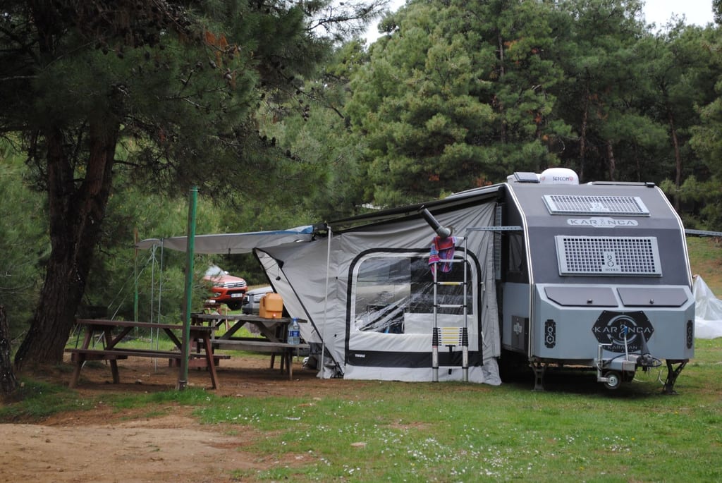 Gray camper trailer with awning set up in a wooded campsite.