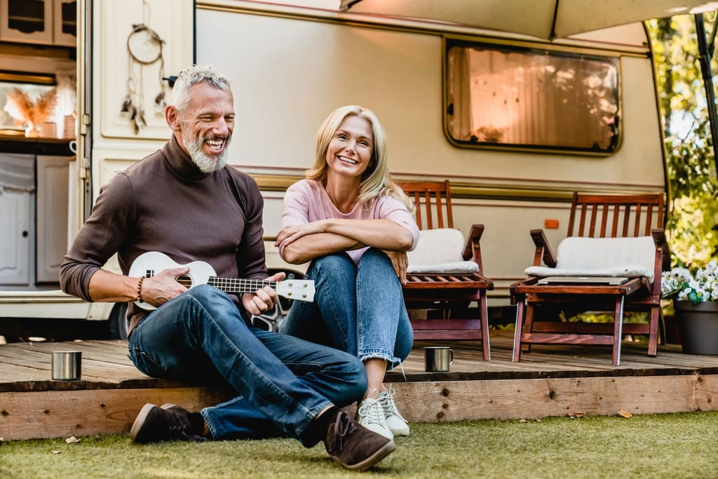A smiling couple relaxing outdoors beside their RV, one of them playing a guitar.